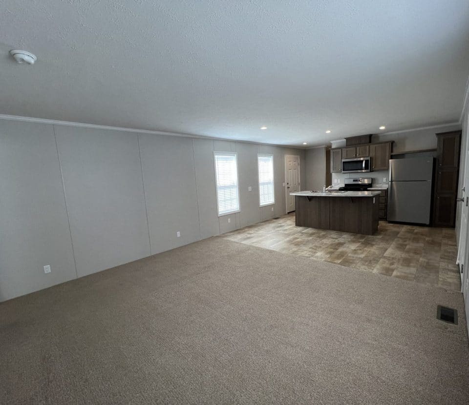 Open-plan room with beige carpet, neutral walls, and a kitchen area with tiled flooring, stainless steel appliances, and an island. Three large windows let in natural light. Room is unfurnished.