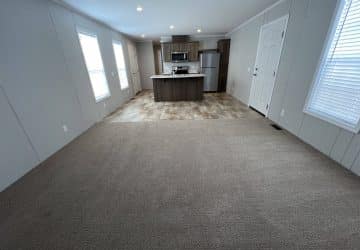 Wide view of an empty, modern open-plan room with beige carpet leading to a kitchen area with tiled flooring, an island, stainless steel appliances, windows, and white walls.