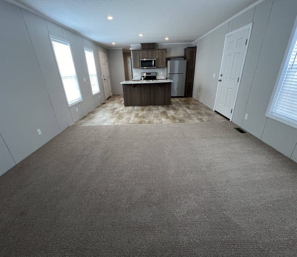 Wide view of an empty, modern open-plan room with beige carpet leading to a kitchen area with tiled flooring, an island, stainless steel appliances, windows, and white walls.
