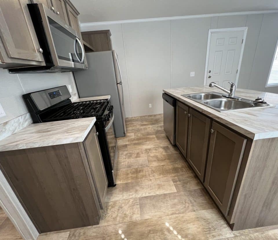 Modern kitchen with wood cabinets, stainless steel appliances, a gas stove, refrigerator, double sink on an island, and light-colored tile flooring. A door and window are visible in the background.