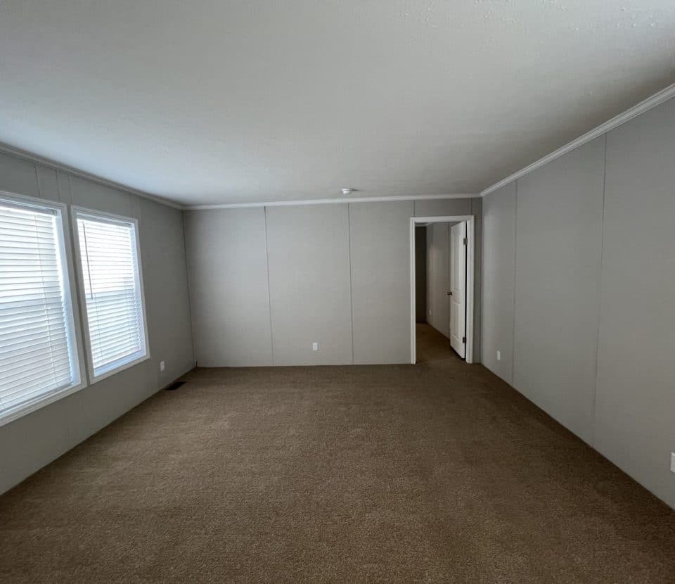 An empty room with beige carpet, light gray walls, two large windows with white blinds on the left, and a doorway leading to another room in the back. The room is unfurnished and well-lit with natural light.