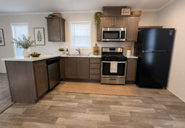 Modern kitchen with brown cabinets, stainless steel appliances, a black refrigerator, white countertops, decorative plants, and wall art. Natural light enters through two small windows above the sink and counter.