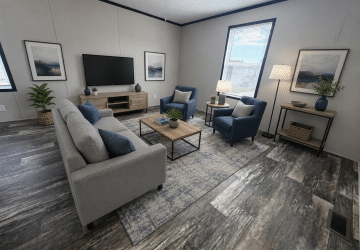 Modern living room with a gray sofa, two blue armchairs, a wooden coffee table, TV on a stand, two framed art pieces, floor lamp, side table with plants, and large windows letting in natural light.