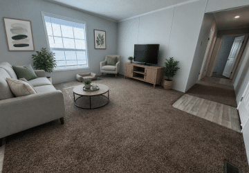 A bright living room with a large window, light gray sofa, a matching armchair, coffee table, TV on a wooden stand, green plants, and neutral wall art. The carpet is brown, and the hallway has light wood flooring.