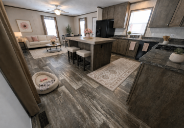 Open-concept kitchen and living room with wood flooring, an island with two stools, dark wood cabinets, a beige sofa, coffee table, and a small dog bed with a toy on the floor. Pink decor and flowers add warmth.