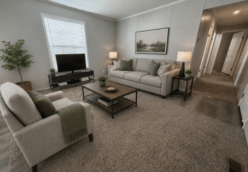 A modern living room with a gray sofa, matching armchair, coffee table, TV on a stand, two lamps, a potted plant, and a landscape painting on the wall. Carpeted floor and a hallway leading to other rooms.