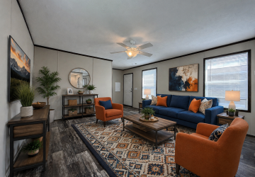 A modern living room with a blue sofa, two orange armchairs, a patterned rug, wooden tables, decorative plants, and wall art. Large windows let in natural light, and a ceiling fan is centered above.