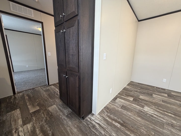 A room with wood-look flooring, beige walls, and dark wood cabinets; an open doorway leads to a carpeted room in the background. The space appears clean and empty.