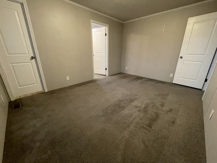 Empty room with beige walls, brown carpet, and three white doors; one door is open, showing a hallway with similar coloring. The room is unfurnished and well-lit by natural light.