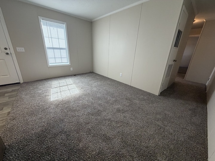 A small, empty room with beige walls, gray carpet, a window with blinds letting in sunlight, and a visible hallway leading to other rooms. The door and part of a vinyl floor are also visible.