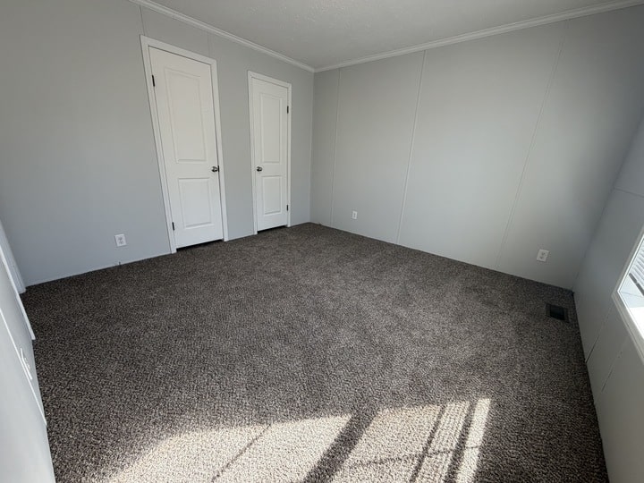 Empty room with light gray walls, two closed white doors, a window letting in natural light, and brown carpet flooring. The space appears clean and unfurnished.