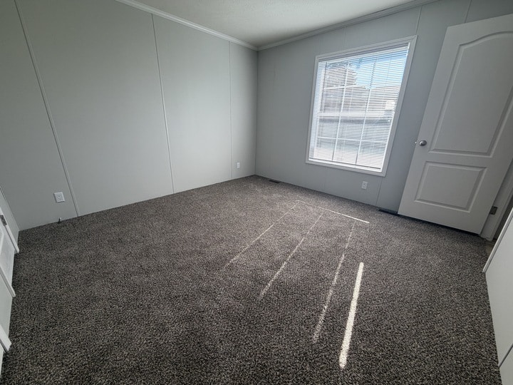 A bright, empty room with light gray walls, a large window with blinds letting sunlight in, and gray carpeted flooring. A white door is partially open on the right side of the room.
