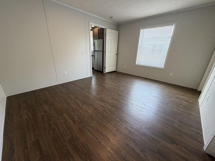 Empty room with dark wood flooring, white walls, a large window, and an open door leading to a kitchen area with visible appliances. Natural light fills the space.