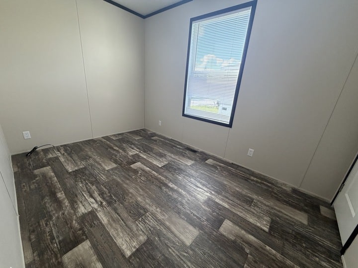 An empty room with light beige walls, dark wood-style flooring, a large window with blinds, and a white door. Sunlight is streaming in through the window.