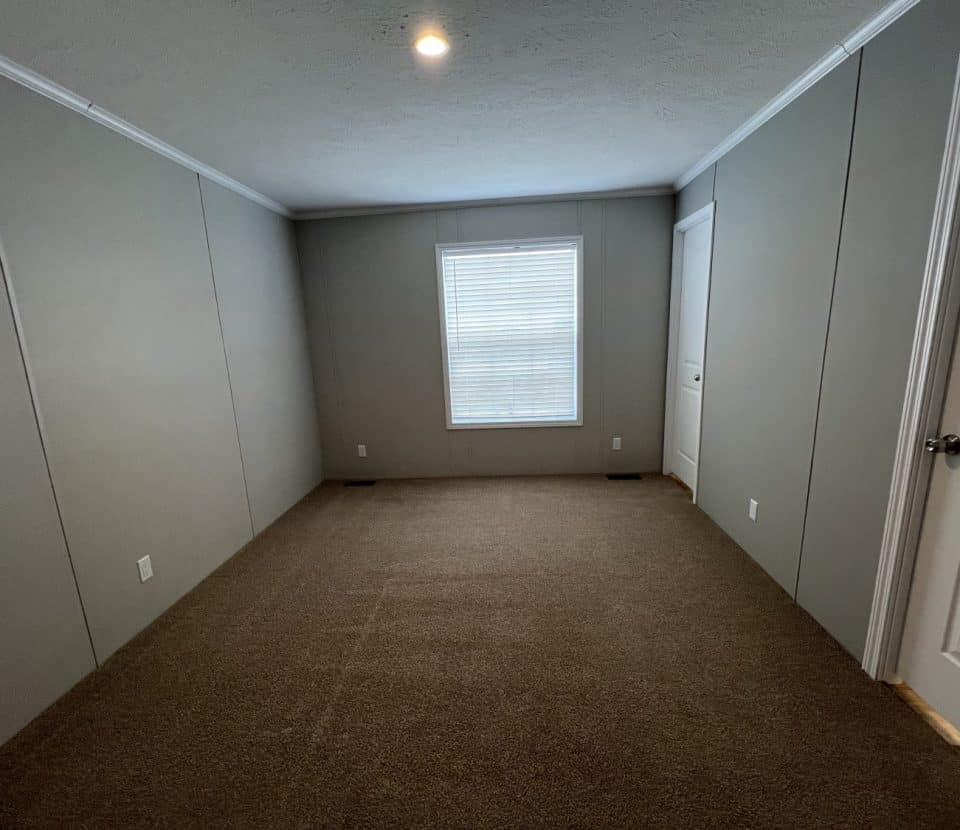 A small, empty room with gray walls, brown carpet, a single window with closed blinds, and two white doors, one on each side of the room. A single ceiling light is turned on.