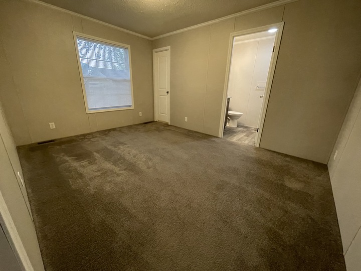 A mostly empty room with beige carpet, beige walls, a window with blinds, two white doors, and an open doorway leading to a bathroom with visible toilet and tile flooring.