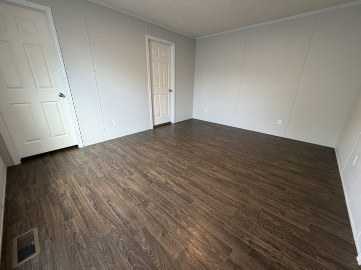 Empty room with light gray walls, dark wood-style flooring, two white doors, and white trim. The room is well-lit and has electrical outlets and a floor vent visible.