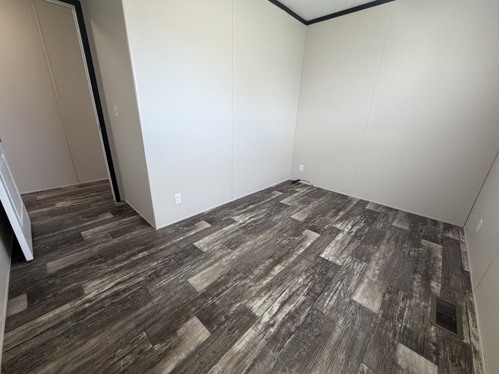 Empty room with light beige walls, dark wood-look vinyl flooring, and a small vent on the floor near the wall. There is an open doorway leading to another room with similar flooring.