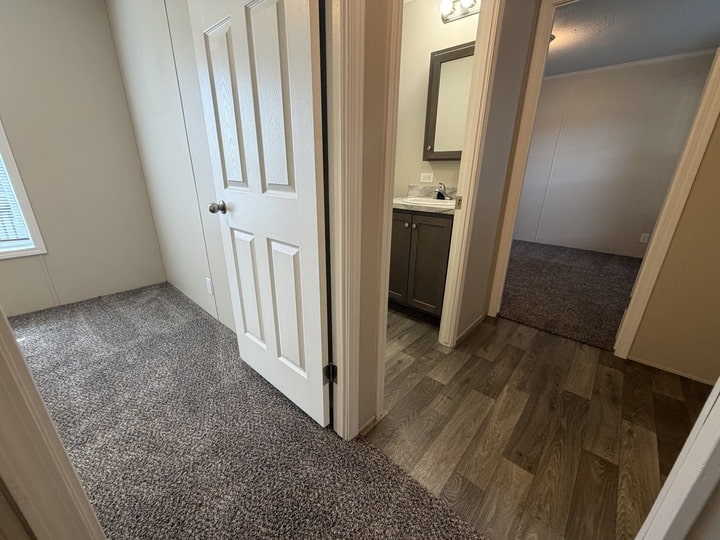 A hallway with carpeted and wood-patterned floors leads to a closed door on the left, a bathroom with a sink and mirror straight ahead, and a carpeted bedroom on the right.