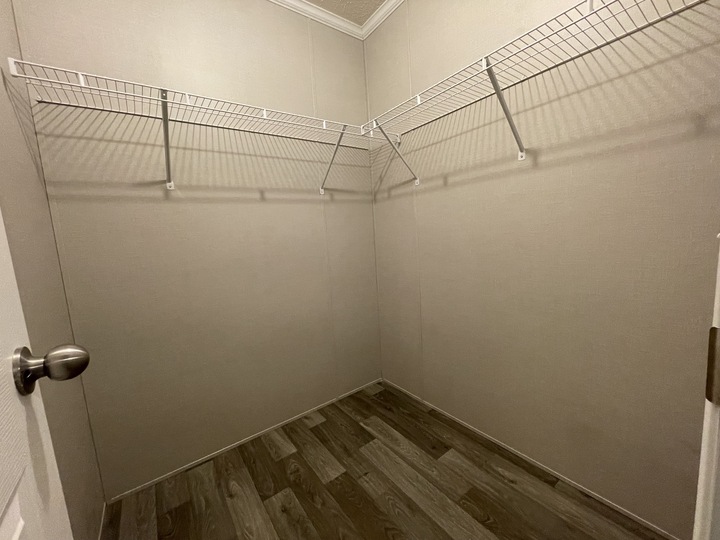 Empty walk-in closet with beige walls, wood-look flooring, and two white wire shelves mounted on adjacent walls. The closet door is open, with a silver door handle visible on the left.