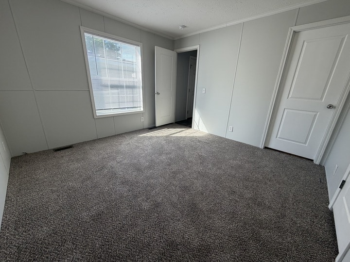 Empty room with light gray walls, a single window with blinds letting in sunlight, two closed white doors, and gray carpeted floor. The space is clean, well-lit, and appears to be a bedroom.