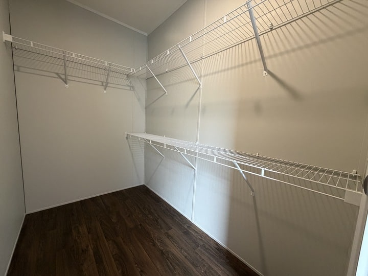 Empty walk-in closet with white wire shelves on two walls, light gray walls, and dark wood flooring. The closet is well-lit and spacious.