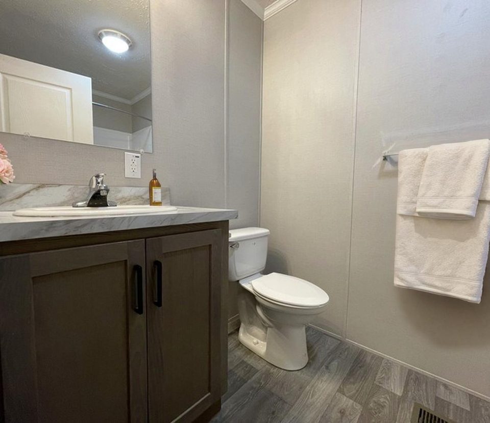A modern bathroom with gray walls and floor, a wooden vanity with a sink, a mirror above, a toilet, a glass shelf with folded white towels, and a bottle of soap. Pink flowers in a vase sit on the counter.