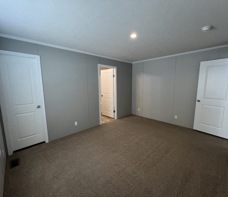 Empty room with beige carpet, light gray walls, white trim, and three closed white doors; one door is open revealing another room. A window with blinds is on the left wall. Ceiling light and smoke detector are visible.