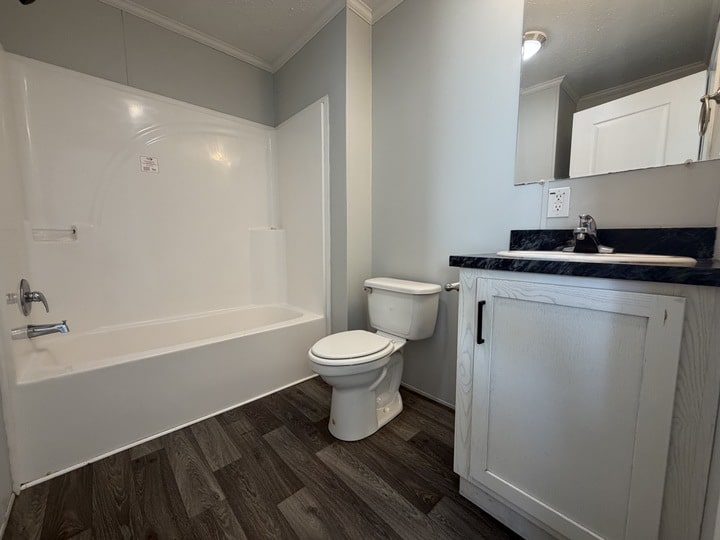 A bathroom with a white bathtub-shower combo, a toilet, and a vanity with a black countertop, sink, and mirror. The floor has dark wood-style planks and the walls are light-colored.