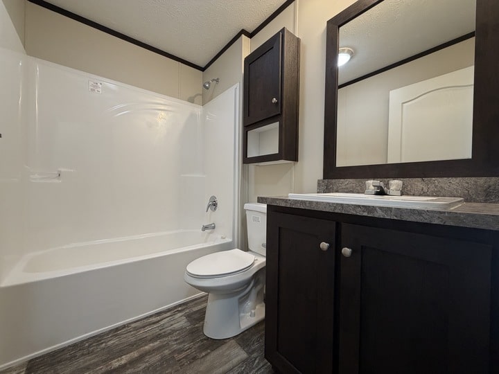 Modern bathroom with a white bathtub-shower combo, a toilet, dark wood cabinets, a large mirror, and gray stone countertop. The floor has a wood-look finish. The room is well-lit and tidy.