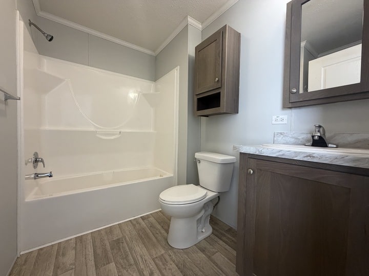 A modern bathroom with a white bathtub and shower, toilet, and wooden cabinets above and below a marble countertop sink. The floor has wood-style tiles and walls are light gray.