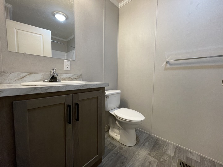 A simple bathroom with a sink and cabinet, a wall-mounted mirror, a toilet, a towel bar, and gray wood-look flooring. The walls are light-colored and plain, with a white door visible in the mirrors reflection.