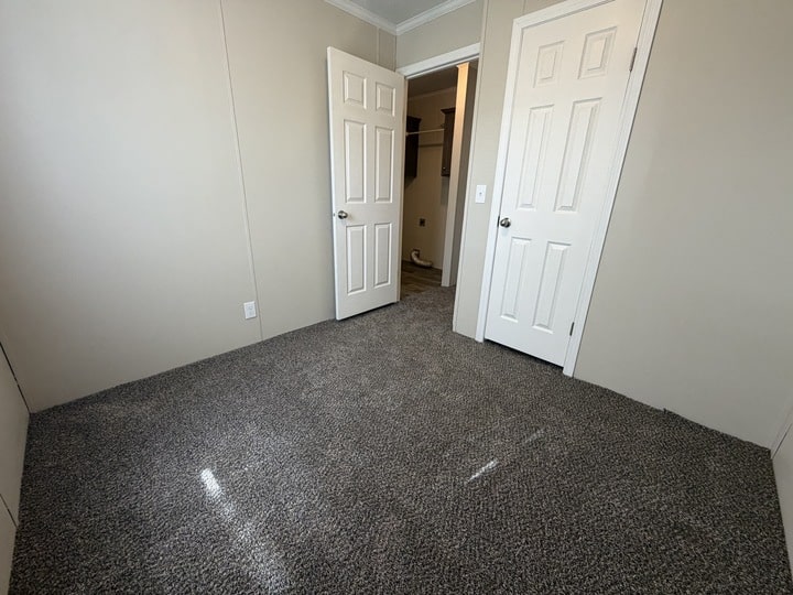 Small, empty room with gray carpet, beige walls, and two white doors—one open, showing a hallway, and one closed. Natural light shines on the carpet from the left side.