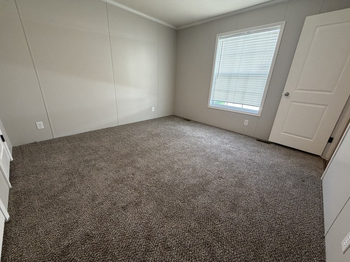 Empty room with beige carpet, light gray walls, a white door slightly open, and a large window with closed blinds letting in some natural light. The space appears clean and unfurnished.