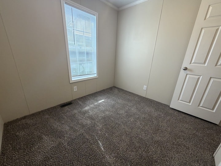Small, empty room with beige walls, a closed white door, a window with blinds letting in sunlight, and gray carpeted flooring.