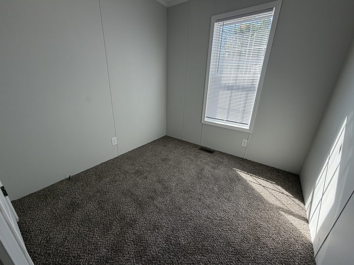 A small, empty room with light gray walls, a window with blinds letting in sunlight, and a brown carpeted floor. The space appears clean and unfurnished.
