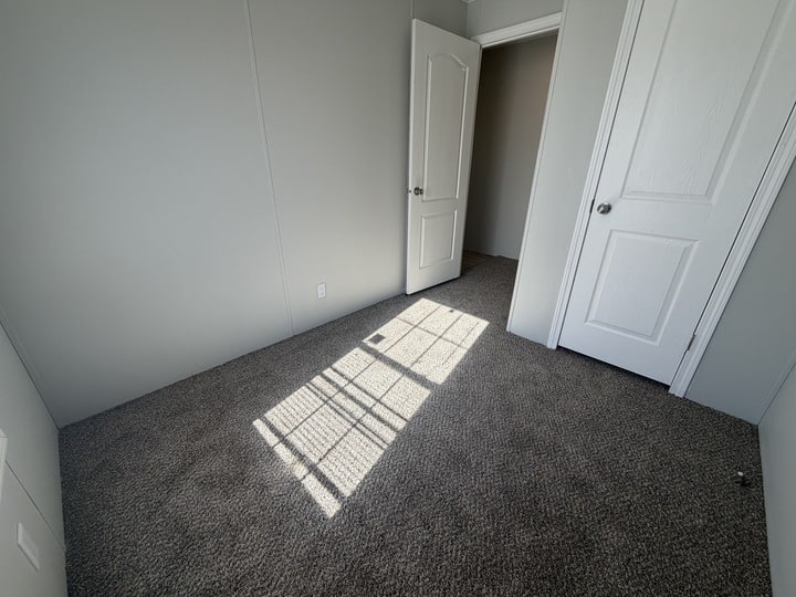 Small, empty room with light gray walls and brown carpet. Sunlight streams through a window, casting shadows on the floor. An open door leads to another room, and a closed white door is on the right.