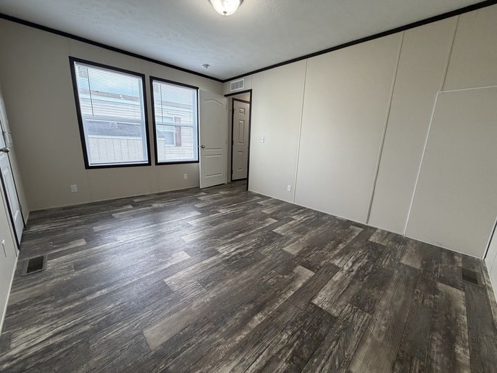 Empty room with wood-patterned vinyl flooring, beige walls, two large windows, a white ceiling, and a ceiling light fixture. A closed door and a small vent are visible on one wall.