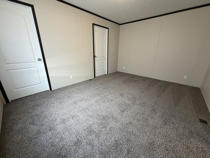 Empty room with beige walls, gray carpet flooring, white baseboards, and two white doors. The room is well-lit and has a simple, clean appearance.