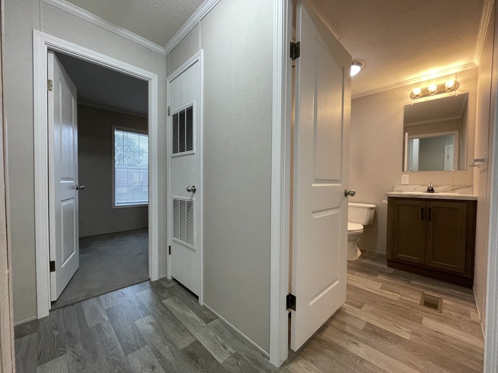 A hallway with light wood flooring, featuring an open door to a carpeted bedroom on the left and an open door to a bathroom with a vanity, mirror, and toilet on the right. White walls and doors throughout.