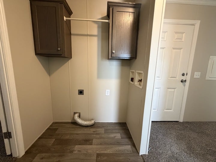 Laundry area with wood cabinets, a hanging rod, wall hookups for washer and dryer, a flexible vent hose on the floor, and a door with a doorknob and lock to the right. The floor is wood and carpeted at the doorway.