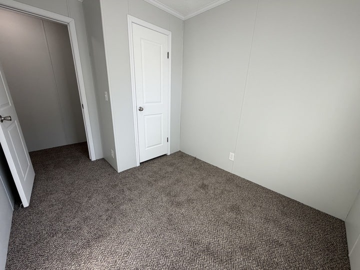 A small, empty room with light gray walls, brown carpet, white trim, and two white doors—one open and one closed. The room appears clean and well-lit.