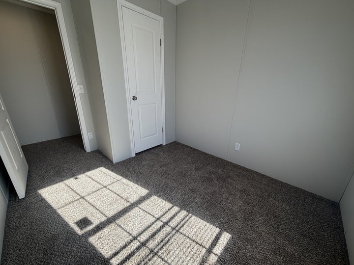 Small empty room with gray carpet, light gray walls, a closed white door, and sunlight streaming through a window, casting shadows on the floor. Another open door leads to an adjacent room.