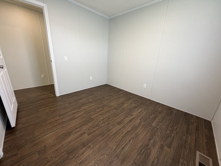 Empty room with light-colored walls, dark wood flooring, white trim, an open doorway on the left, and electrical outlets visible on the walls. A floor vent is in the lower right corner.