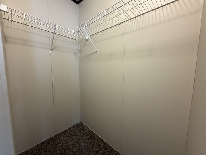 Empty walk-in closet with beige walls, brown carpeted floor, and two white wire shelves running along the upper sections of two walls.