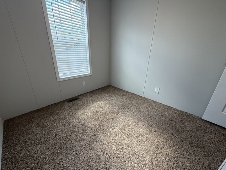 Empty room with beige carpet, light gray walls, a closed white door, and a window with white blinds letting in natural light.