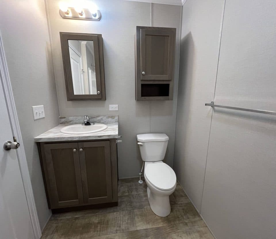 A small, modern bathroom with a sink and vanity, a wall-mounted mirror cabinet above the sink, a toilet, an overhead storage cabinet, a towel bar, and neutral-colored walls and floor tiles.
