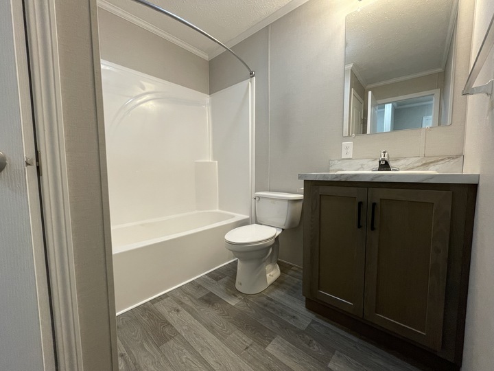 Modern bathroom with a white bathtub and shower, a toilet, and a dark wood vanity with a marble countertop and mirror above. The floor has gray wood-style vinyl planks, and the walls are light-colored.