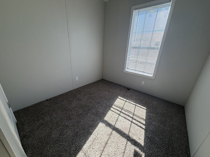 A small, empty room with gray carpet, plain white walls, and a single window with blinds letting in sunlight and casting shadows on the floor.