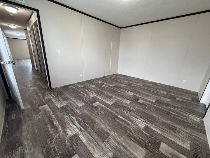 Empty room with light beige walls, dark trim, and wood-patterned vinyl flooring. Doorways lead to a hallway with similar flooring, and there are multiple electrical outlets on the walls.